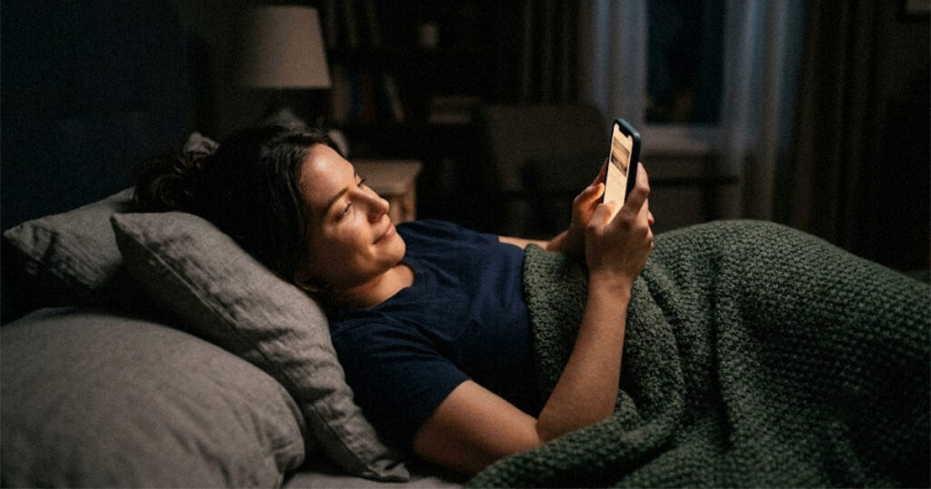 A person reading on a smartphone in a dark bedroom with a soft, steady screen glow on their face